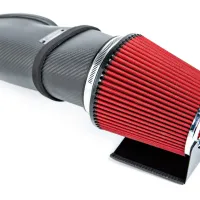 A red and black cone-shaped air filter attached to a carbon fiber intake system, resting on a white surface.