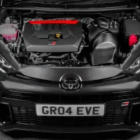 Car engine bay containing a visible engine with red and black components, and a front license plate reading "GR04 EVE", set within a black Toyota vehicle in an indoor setting.