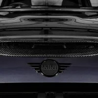 A carbon fiber hood scoop and "MINI" emblem on a sleek, dark-colored surface, reflecting ceiling patterns, likely part of a Mini Cooper car's exterior.