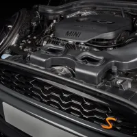Car engine labeled "MINI," featuring a prominent airflow system and various mechanical components, situated within the open hood of a vehicle with a visible front grille and an orange "S" badge.