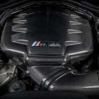 A carbon fiber engine cover labeled "M Power" sits at the center of a car engine bay, with various components and wires surrounding it, signifying a high-performance vehicle.