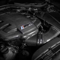 Carbon fiber engine cover with a BMW M Power logo on a high-performance car engine in a dark, industrial garage setting.