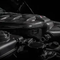 Carbon fiber engine cover with Eventuri branding sits in a dimly lit car engine compartment with various mechanical components surrounding it. Text on the cover reads "Eventuri, B58 Carbon Plenum."