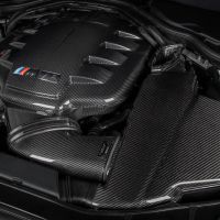 Carbon fiber engine cover labelled "M5" with colored stripes in a car engine bay; visible surrounding components include various cables and other engine parts.