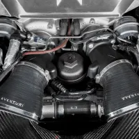 Engine components with "Eventuri" branded air intakes positioned centrally, surrounded by various cables and metal parts inside the car engine bay. The air intakes are covered by patterned carbon fiber material.