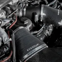Engine compartment displaying "Eventuri” air intake system with carbon fiber tubing, situated among metal and rubber pipes, various fasteners, and components. Engraved text on metal reads “0603C10105.”