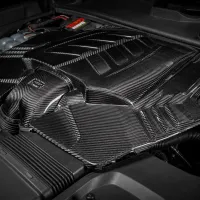 A carbon fiber engine cover sits within a car's engine bay, showcasing sleek design elements. Text reads, "I3-LASNIKO Transfomotive."