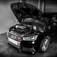 A black car with its hood open, showcasing the engine, is stationary in a dimly lit garage next to a white brick wall. Text on the license plate reads "EVENTURI".