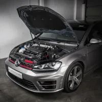 A silver Volkswagen car with its hood open, revealing the engine, parked inside a garage with white brick walls. The front license plate reads "VENTURI."