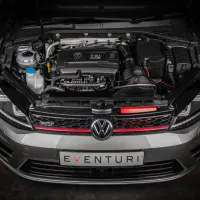 The engine compartment of a silver Volkswagen GTI is shown with the hood open, featuring a TSI engine. A white license plate reads "EVENTURI," and the car is in a dimly lit garage.