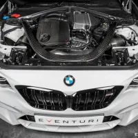 A BMW car's engine bay with a carbon fiber intake system, displayed in a garage with the hood open. The front grille features the BMW logo. The front plate reads "EVENTURI."