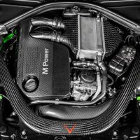 A carbon fiber engine cover labeled "M Power" sits between black hoses and metal components, surrounded by neon green car chassis and black wiring inside a vehicle's engine bay.