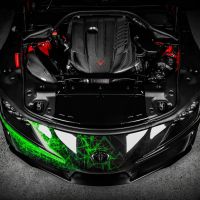 A car's engine bay with a detailed, carbon-fiber engine cover is displayed on a sleek black car, highlighted with green neon graphical patterns, situated on a concrete floor.