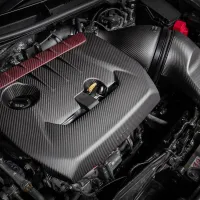 A carbon fiber engine cover sits atop a car engine, featuring sleek design and precise detailing in a workshop setting. Nearby components and wiring are visible, highlighting a well-maintained automotive system.