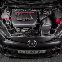 The opened hood reveals a well-kept car engine, with visible carbon fiber components, and "GR04 EVE" on the license plate, set in a workshop with a concrete floor.