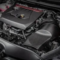 Car engine covered with a sleek, black carbon fiber cover; surrounded by various hoses, wires, and coolant tank, within a Toyota vehicle's engine compartment.
