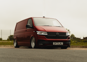 A red Volkswagen van with license plate "GV73 LNJ" is parked on a road with a grassy area and buildings in the background under an overcast sky.