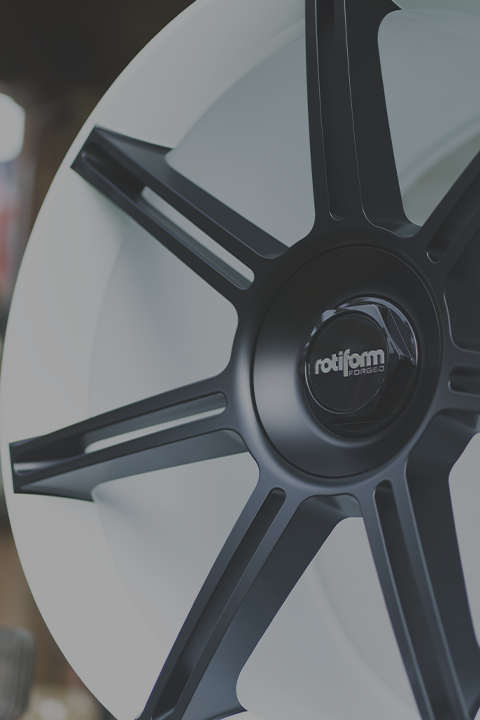 A black, multi-spoke alloy wheel with "rotiform FORGED" on the center cap leans against a white surface in a dimly lit workshop.