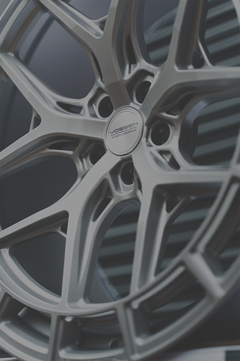 A metallic alloy wheel is stationary with a modern, intricate design; the Vossen logo is visible in the center. The background appears to be a dark, striped surface. Text reads: "VOSSEN HYBRID FORGED."