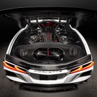 A white Corvette showcases its powerful engine under an open hood in a dimly lit garage. Text reads "CORVETTE" on the rear.