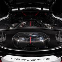 A Corvette with its rear engine compartment open, showcasing a carbon fiber engine component with a red stripe. The surrounding area features sleek, black detailing in a brightly lit environment. "CORVETTE" text is visible at the bottom.