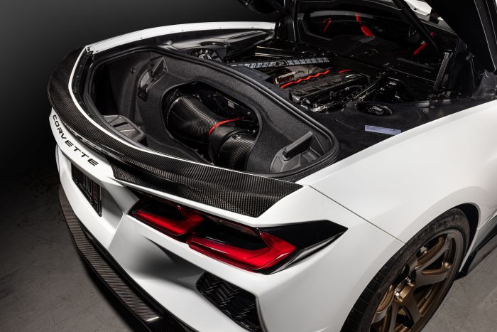 A white car with the rear hood open revealing a detailed engine compartment, featuring carbon fiber and red accents, in a studio setting. "CORVETTE" text is visible on the rear bumper.