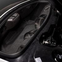 Car trunk with black carbon fiber and gray interior, showing components and storage space. Surrounding the context is the interior of the car, partly showing an engine.
