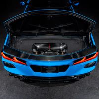 A blue sports car with its rear trunk open, revealing a carbon fiber component, is parked on a dark surface in a low-lit environment.