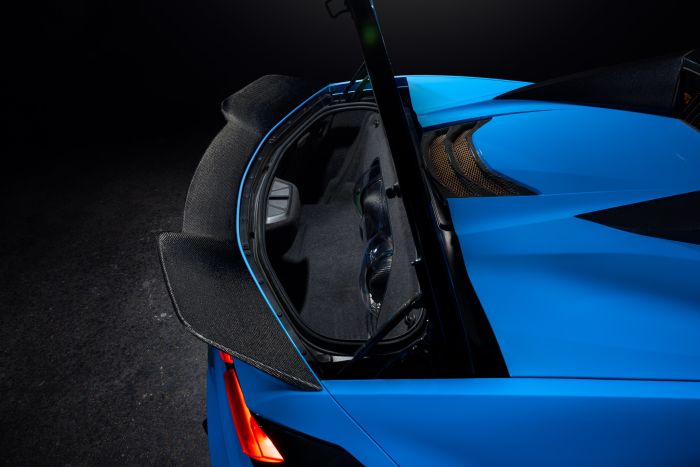 A blue sports car's rear hatchback is open, revealing a spacious trunk with carbon fiber detailing, under dark ambient lighting. The left rear taillight is visible.