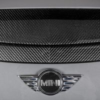 A carbon fiber air vent is mounted on a metallic surface, with the "MINI" emblem featuring wings situated below it. The metallic surface appears to be part of a vehicle.