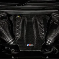 A carbon fiber engine cover, displaying a red, blue, and light blue-striped "M" logo, is situated within a car's polished engine bay.