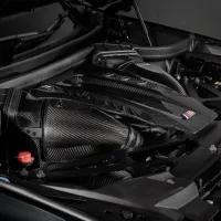 Carbon fiber car engine cover on a high-performance engine, featuring a blue, violet, and red "M" logo, set within a dark mechanical environment.
