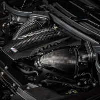 Carbon fiber engine cover and air intakes, all adorned with a small colored emblem, situated in the sleek engine bay of a modern car.