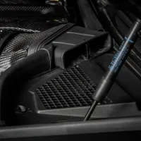 Carbon fiber intake system connected to the air filter housing, situated within the hood compartment of a vehicle. The compartment features various black components and a hydraulic rod labeled "Stabilus."