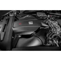 A black, carbon fiber car engine with complex tubing and a red arrow symbol is mounted inside the engine bay of a vehicle.