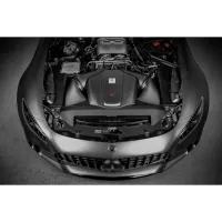A car with an open hood showing its engine compartment, in a dimly lit garage. The car has a prominent grille featuring a Mercedes-Benz logo.