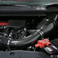 Carbon fiber air intake system installed in a car engine bay, with red components and a visible battery, surroundings include tubes and other engine parts. Text reads: "EVENTURI."