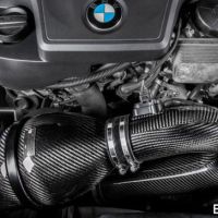 A high-performance car engine featuring a carbon fiber air intake system with metal clamps. The textured carbon fiber contrasts with the polished engine components. Text: "EVENTURI" at the bottom right.