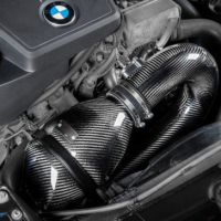 Engine compartment featuring a BMW engine with a carbon fiber intake system by Eventuri, installed and surrounded by various automotive components within a car’s engine bay. Text: “EVENTURI.”
