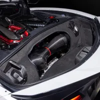 Car engine with carbon fiber components is in a trunk compartment, surrounded by white bodywork, parked on a dark surface. The engine comprises black and red elements with intricate detailing.