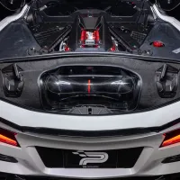 The rear engine compartment of a white sports car displays a high-performance engine with red accents and carbon fiber details under a clear cover, set against a dark background.
