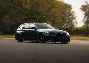 A sleek, black BMW wagon is parked on an empty road, surrounded by tall green trees on a cloudy day.