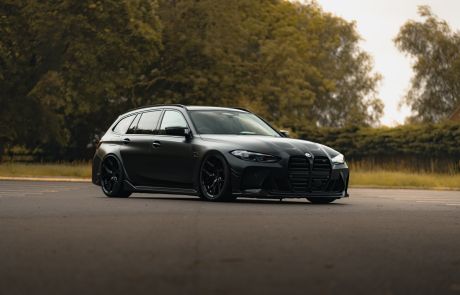 A sleek, black BMW wagon is parked on an empty road, surrounded by tall green trees on a cloudy day.