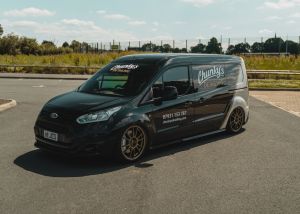 A black van parked on an asphalt surface with "Chunky's Detailing," a contact number, and website (chunkysdetailing.com) displayed on its side. Background includes greenery, fencing, and a clear sky with clouds.
