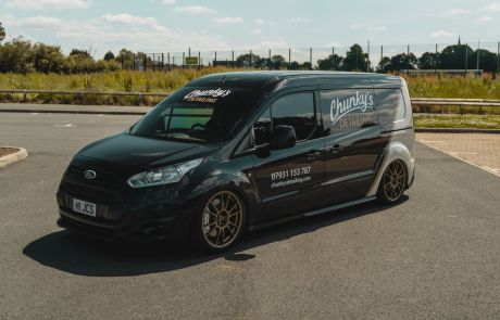 A black van parked on an asphalt surface with "Chunky's Detailing," a contact number, and website (chunkysdetailing.com) displayed on its side. Background includes greenery, fencing, and a clear sky with clouds.