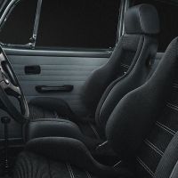 The image features the interior of a car, with black, sporty Recaro seats and a steering wheel. The setting is the dark interior cabin, highlighting vintage automotive design.