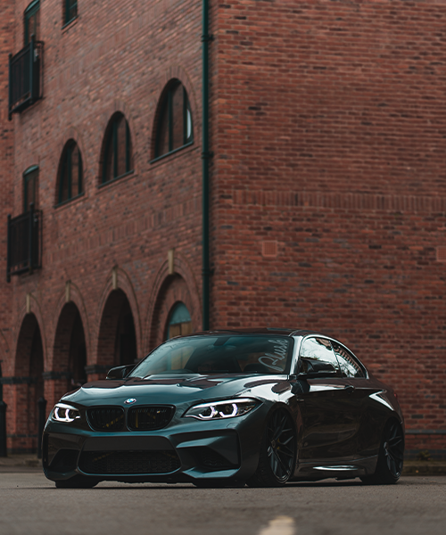 A sleek, black BMW car is parked on a street, set against a backdrop of red brick buildings with arched windows and balconies.