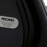 A close-up of a black car seat features a metallic plaque that reads: "RECARO Sportster CS Exclusive Edition Nürburgring No. 1 of 999." Red stitching and background are partially visible.