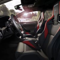 A luxurious car interior featuring black and red Recaro racing seats, a modern dashboard, and a steering wheel with red stitching. The car is on a winding forest road, seen through the windshield. Text on seats reads: "RECARO" and "Nürburgring".