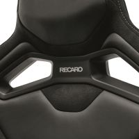 A black, high-backed Recaro car seat with two rectangular cutouts and "RECARO" logo centrally positioned, with detailed stitching visible on the edges.
